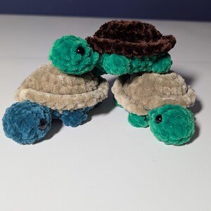 May include: Three handmade crochet turtles in shades of green, brown, and tan. The turtles have textured shells and heads, with black eyes. The turtles are stacked on top of each other.