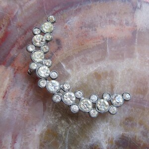 May include: A crescent-shaped ear cuff crafted from dark metal, featuring a cluster of round, clear, faceted rhinestones of varying sizes arranged in a dense, organic pattern that follows the curve of the piece.