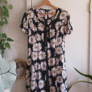 May include: A vintage midi dress with a daisy floral pattern in black and beige. The dress features short sleeves, a tie neckline, and a polka dot panel down the front. It hangs on a wooden hanger against a white door.
