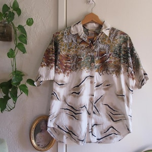 May include: A white button-up shirt with a short sleeve and a safari print featuring zebras, leopards, and other animals. The shirt has a brown, green, and white color scheme.