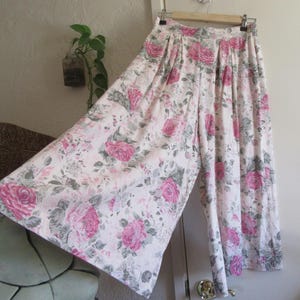 May include: Wide-leg pants with a floral rose pattern in pink, white, and gray. The pants are hanging on a wooden hanger. The fabric is lightweight and appears to be cotton or a similar material. The pants are pleated at the waist.