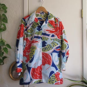 May include: A long-sleeved button-down shirt with a vibrant, abstract pattern. The shirt features a mix of red, blue, green, and white shapes. It hangs on a wooden hanger against a white door.
