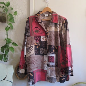 May include: A long-sleeved button-down shirt with a patchwork design. The shirt features a mix of colors including red, brown, black, and cream. The pattern includes floral and geometric shapes. The shirt is hanging on a wooden hanger.