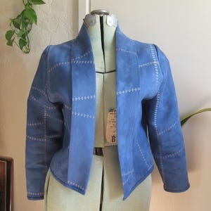 May include: A light blue suede jacket with white stitching details. The jacket has a unique patchwork design and a collarless, open front. The sleeves are long, and the jacket is displayed on a dress form.