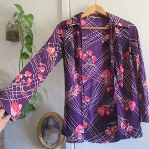 May include: A vintage purple button-up shirt with a floral pattern. The shirt features a design of pink flowers and white lines. The long-sleeved shirt is hanging on a wooden hanger.