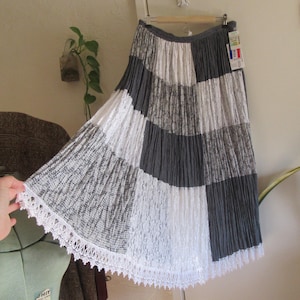 May include: A patchwork midi skirt in shades of gray and white, with a scalloped lace trim. The skirt is hanging on a wooden hanger. A tag with French text is attached to the skirt.