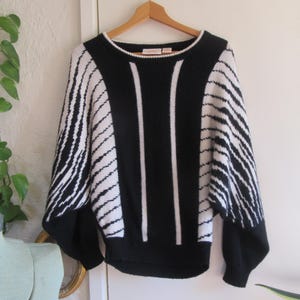 May include: A black and white sweater with a unique design. The sweater features a black base with white vertical stripes down the front and white diagonal stripes on the sleeves. The sweater is hanging on a wooden hanger.