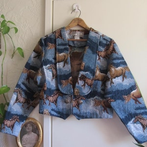 May include: A cropped jacket featuring a woven tapestry pattern of brown horses running across a blue and white cloudy sky background. The jacket has a wide shawl collar, long sleeves, and two metallic buttons at the front closure.