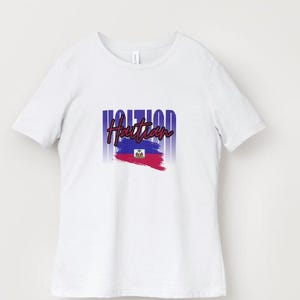 Pu&ograve; includere: T-shirt bianca con la scritta "Haitian" in un font stilizzato sopra una grafica della bandiera haitiana. Il design della bandiera include pennellate blu e rosse e lo stemma haitiano. La maglietta ha un girocollo e maniche corte.