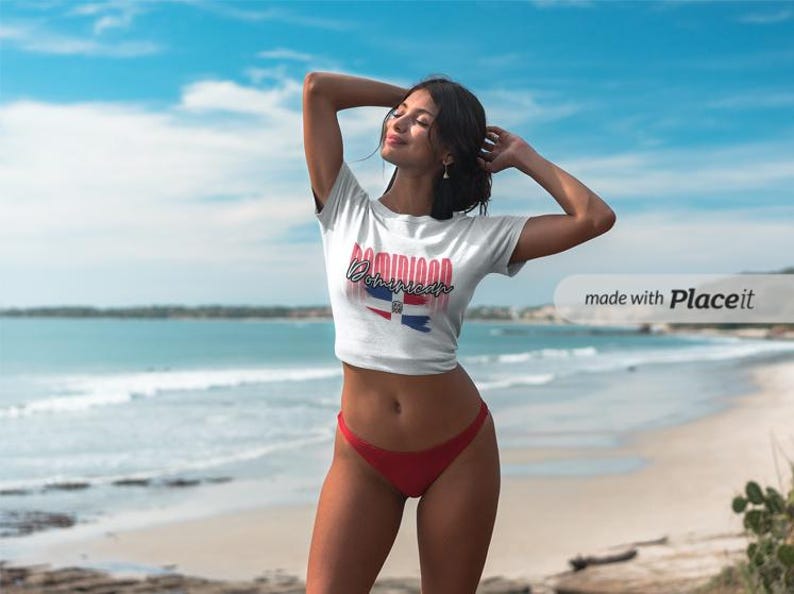 Pu&ograve; includere: Un top corto bianco con la scritta "Dominican" in rosso e blu, insieme a un disegno della bandiera. La modella indossa slip rossi ed &egrave; su una spiaggia con cielo blu.