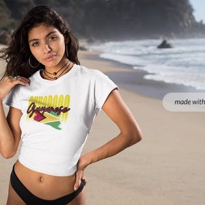 Pu&ograve; includere: T-shirt bianca con la scritta "Guyanese" in giallo e rosso, e la bandiera della Guyana. La maglietta &egrave; indossata da una persona sulla spiaggia.