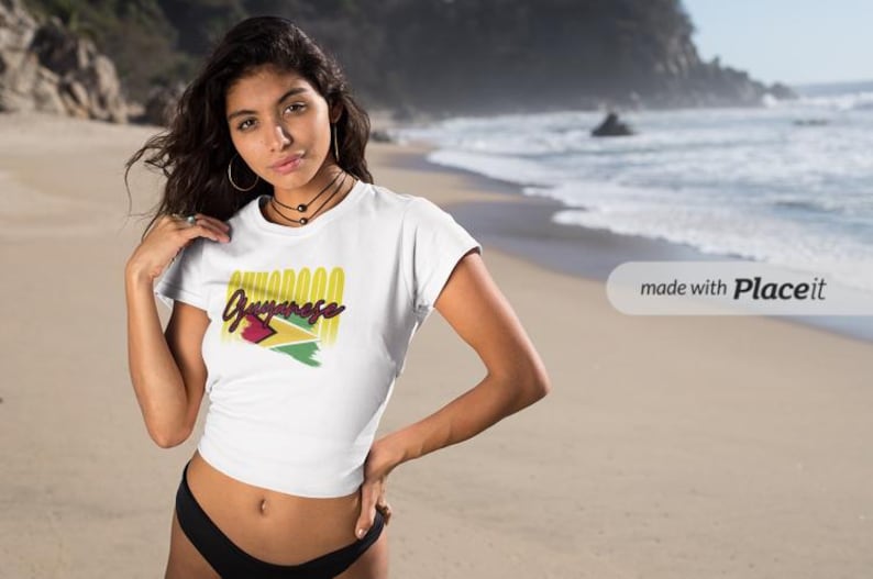 Pu&ograve; includere: T-shirt bianca con la scritta "Guyanese" in giallo e rosso, e la bandiera della Guyana. La maglietta &egrave; indossata da una persona sulla spiaggia.