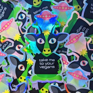 May include: A holographic sticker featuring a cartoon cow wearing a black shirt with the text "Take me to your vegans". The cow is surrounded by other holographic stickers with alien spaceships and the text "Take me".