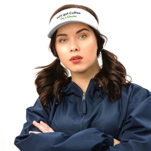 May include: White visor with the text "Let's get Coffee On a Cruise" in green and black. The visor is worn by a person wearing a navy blue jacket.