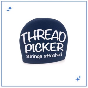 May include: A navy blue thread picker with white text that reads "THREAD PICKER strings attached." The thread picker is a small, rounded shape, and the text is in a handwritten style.