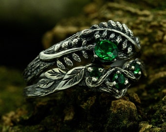 May include: A silver ring with a leafy design and green gemstones. The ring is set on a mossy background.