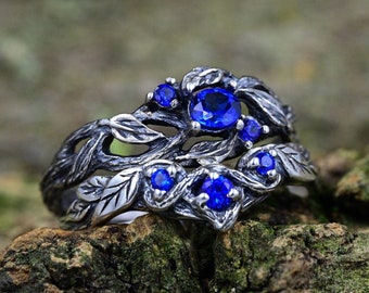May include: A silver ring with a blue sapphire gemstone set in a tree branch design. The ring has a delicate, intricate design with multiple leaves and branches.