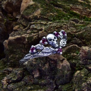 Könnte beinhalten: Ein silberner Ring mit einem Schädel- und Blattmotiv, verziert mit kleinen, runden, roten Edelsteinen. Der Ring ist vor einem strukturierten, moosbewachsenen Hintergrund platziert, der die detaillierte Handwerkskunst hervorhebt.