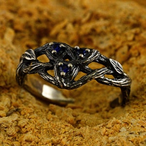 Peut inclure: Une bague en argent avec un motif tissé orné de pierres précieuses bleues. La bague est posée sur un fond brun texturé.