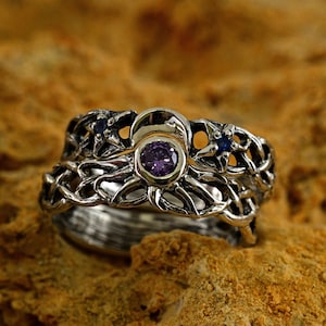 May include: A silver ring with a delicate, intricate design featuring a purple gemstone surrounded by smaller blue gemstones. The ring is set against a textured brown background.