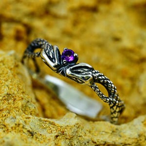 May include: A silver ring with a purple gemstone set in the center. The ring is designed in the shape of two intertwined snakes.