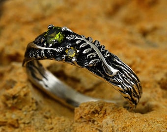 May include: A silver ring with a textured design resembling a tree branch and leaves. The ring features three green gemstones.