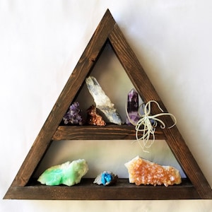以下が含まれることがあります： 2 段の棚が付いた三角形の木製棚に、さまざまなクリスタルと小さなエアプラントが飾られています。クリスタルは、紫、緑、オレンジ、青など、さまざまな色をしています。