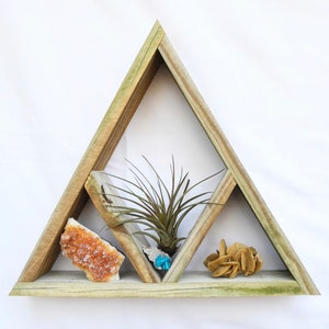 Puede incluir: Una estantería de madera triangular con tres compartimentos. La estantería está decorada con una pequeña planta de aire, un grupo de cristales naranjas y un grupo de cristales marrones.