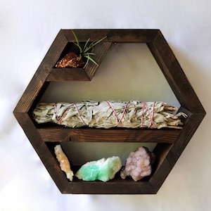 以下が含まれることがあります： 3 段の棚が付いた六角形の棚。一番上の棚には、小さな鉢植えの植物と茶色のクリスタルがあります。真ん中の棚には、白いセージの束があります。一番下の棚には、薄い茶色のクリスタル、緑色のクリスタル、ピンク色のクリスタル、白いクリスタルの 4 つのクリスタルがあります。