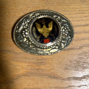 May include: Oval-shaped silver-tone belt buckle with a central emblem featuring a gold eagle on a blue background. The buckle has a decorative border with floral and leaf patterns. The text "ORDER OF THE EAGLE" is visible.