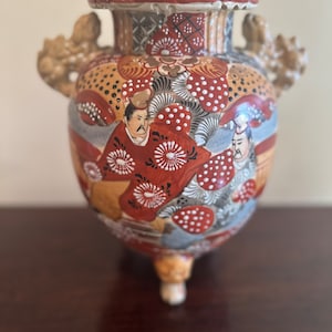 Puede incluir: Un jarrón de cerámica con un cuerpo bulboso y una boca ancha. El jarrón está decorado con un patrón floral colorido y dos figuras con vestimenta tradicional japonesa. El jarrón tiene dos asas en forma de dragón y tres pies.