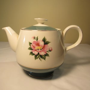 May include: A white teapot with a blue and gold trim and a pink floral design. The teapot has a round body and a handle on the right side.