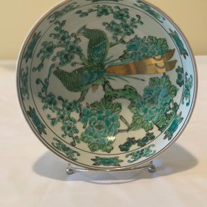 May include: A white ceramic bowl with a teal and gold floral and bird design. The bowl is supported by a silver metal stand. The interior of the bowl features a detailed illustration of birds and flowers.