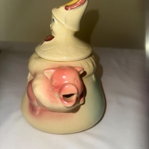 Puede incluir: Una tetera de cerámica vintage con un diseño caprichoso. La tetera es principalmente de color crema, con una boquilla en forma de cerdo y una tapa con una figura que lleva un sombrero. El cerdo es rosa y el sombrero tiene detalles rojos y amarillos.