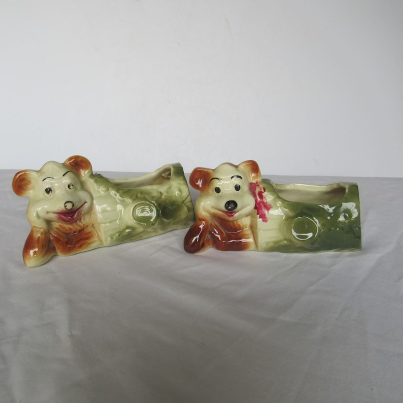 May include: Two ceramic planter pots shaped like a log with a cartoon bear face on each end. The bears are brown and white with black eyes and a red nose. The logs are green and brown with a white background.