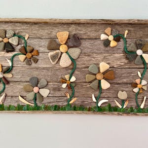May include: A rustic wooden frame with a nature-inspired design featuring multiple flowers made from various stones and pebbles. The flowers are arranged in a row on a wooden background with green string vines.