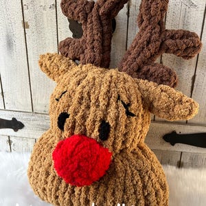 May include: A plush, handmade reindeer head with brown antlers, a tan face, black eyes, and a red nose. The text "stacey made it!" is visible on the bottom left. This festive decoration is perfect for the holiday season.