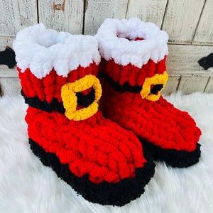 Puede incluir: Un par de pantuflas de ganchillo hasta el tobillo con un cuerpo rojo brillante, un puño blanco grueso y difuso en la parte superior, una banda negra en la base y un detalle de hebilla cuadrada amarilla en el frente, hechas de hilo suave.