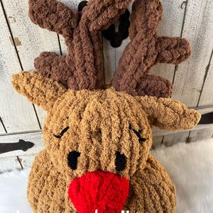 May include: A plush, handmade reindeer head with brown antlers, a tan face, black eyes, and a bright red nose. The text "stacey made it!" is written in white script across the bottom. This festive decoration is perfect for the holiday season.