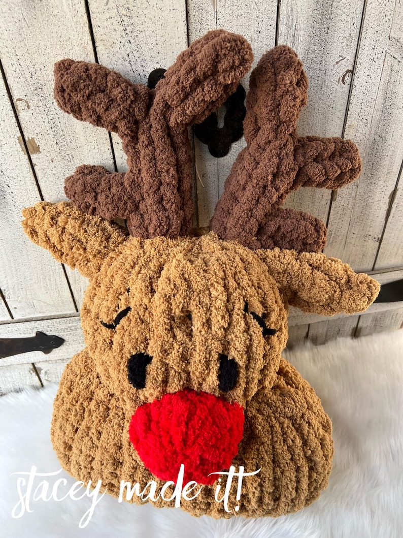 May include: A plush, handmade reindeer head with brown antlers, a tan face, black eyes, and a bright red nose. The text "stacey made it!" is written in white script across the bottom. This festive decoration is perfect for the holiday season.