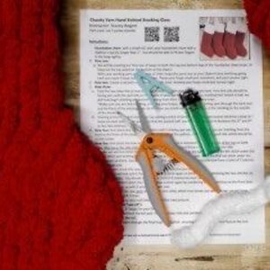 May include: A red knitted stocking with a chunky yarn texture, along with a pair of scissors, a lighter, and white yarn. The image also includes a printed instruction sheet with the title "Chunky Yarn Hand Knitted Stocking Cozy" and a QR code.