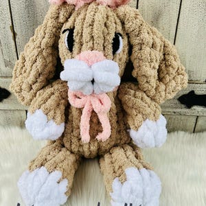 May include: A handmade, plush bunny in shades of tan and white, with a pink bow and accents. The bunny has large, floppy ears and is made of a soft, textured material. The text "stacey made it!" is written in a cursive font at the bottom.