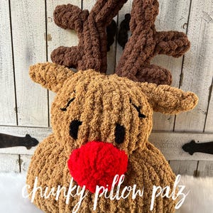 May include: A handmade, plush reindeer pillow with brown antlers, tan body, black eyes, and a red nose. The pillow has the words "chunky pillow palz" and "REINDEER PILLOW TUTORIAL" in white script.