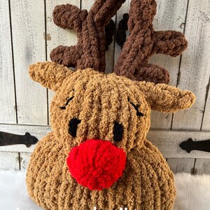 May include: A plush, handmade reindeer pillow with brown antlers, a tan body, black eyes, and a bright red nose. The text "stacey made it!" is written in white script at the bottom. The pillow is designed for holiday decor.