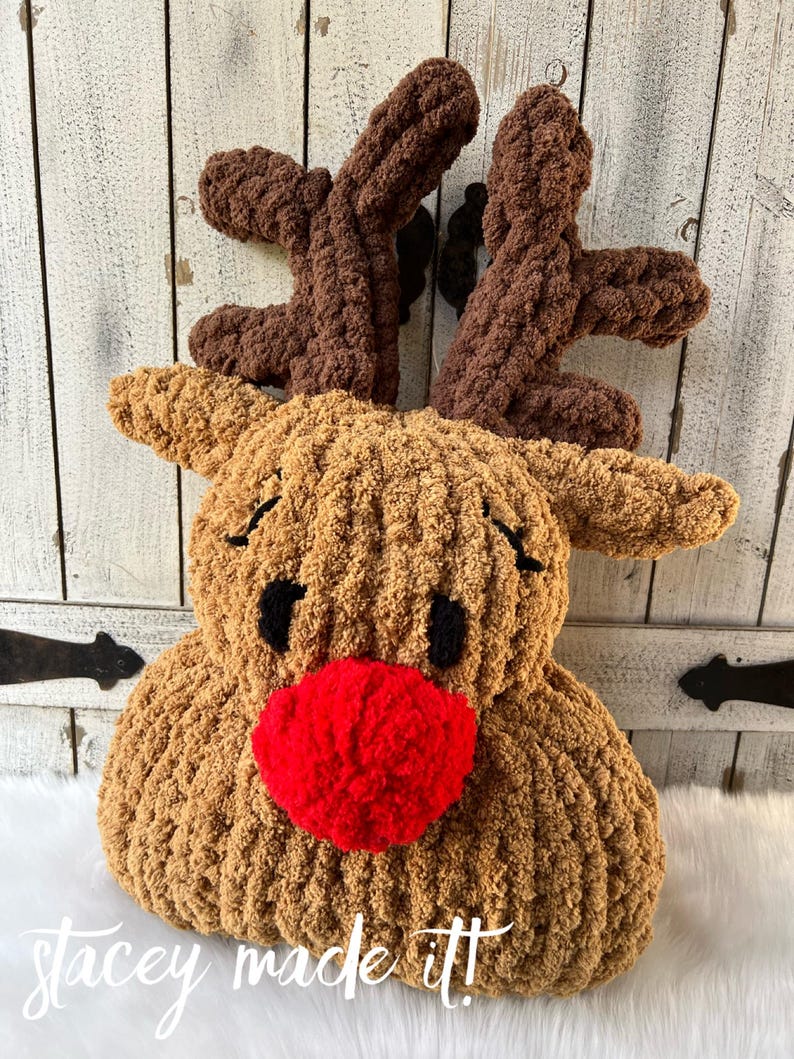 May include: A plush, handmade reindeer pillow with brown antlers, a tan body, black eyes, and a bright red nose. The text "stacey made it!" is written in white script at the bottom. The pillow is designed for holiday decor.