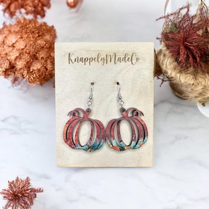 May include: A pair of pumpkin-shaped earrings with a colorful, textured design. The earrings are made of wood and have silver-colored ear wires. The earrings are displayed on a wooden plaque with the text "KnappelyMadeCo".