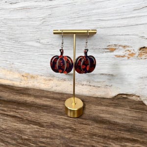May include: A pair of pumpkin-shaped earrings with a dark purple base and red leaf pattern. The earrings are displayed on a gold-colored earring stand against a light wood background. The earrings have silver-colored hooks.