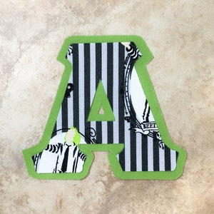 May include: A green felt letter A with a black and white striped fabric background. The fabric has a pattern of a skeleton and a mirror with the word "SHEET" on it.