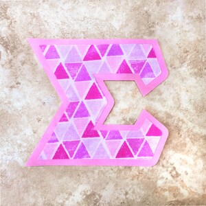 May include: A pink fabric letter "E" with a geometric pattern of pink and purple triangles.