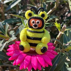 May include: A small, textured yellow and black bee-themed bear figurine with a round orange face and black antennae, sitting atop a vibrant, multi-petaled hot pink flower. The bear has black paws and a black-rimmed hood around its face.